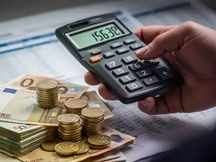 Eine Hand hält einen Taschenrechner über Stapeln von Euro-Banknoten und Münzen, mit einem unscharfen Finanzdokument im Hintergrund. - Foto: über boerse-global.de