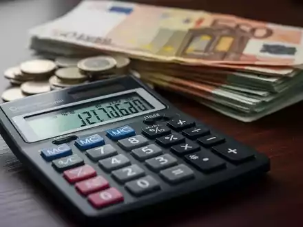 Ein Taschenrechner zeigt Finanzzahlen, mit einem Stapel Euro-Banknoten und Münzen im unscharfen Hintergrund auf einem dunklen Holztisch. - Foto: über boerse-global.de