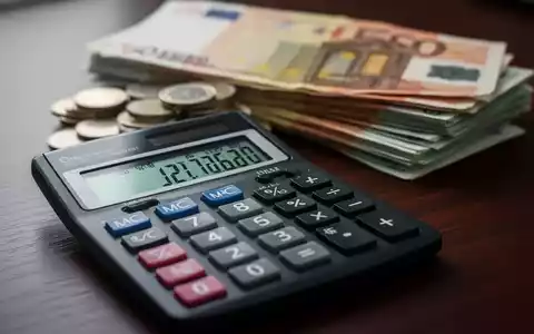 Ein Taschenrechner zeigt Finanzzahlen, mit einem Stapel Euro-Banknoten und Münzen im unscharfen Hintergrund auf einem dunklen Holztisch. - Foto: über boerse-global.de