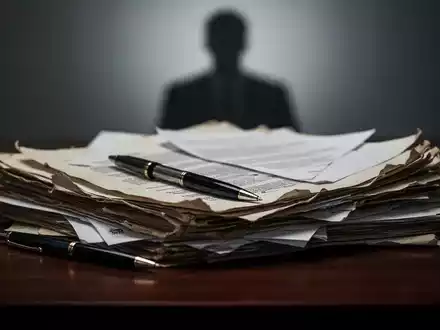 Ein Stapel alter juristischer Dokumente und ein Stift auf einem dunklen Holztisch, im Hintergrund eine unscharfe Silhouette. - Foto: über boerse-global.de