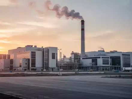 Industriegebiet mit großen Fabrikhallen und einem Schornstein, bei Sonnenuntergang. Ein weites, leeres Gelände im Vordergrund, das zukünftige Entwicklung andeutet. - Foto: über boerse-global.de
