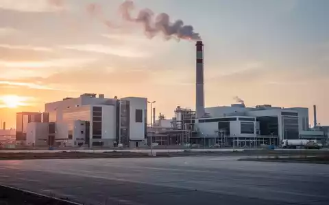 Industriegebiet mit großen Fabrikhallen und einem Schornstein, bei Sonnenuntergang. Ein weites, leeres Gelände im Vordergrund, das zukünftige Entwicklung andeutet. - Foto: über boerse-global.de