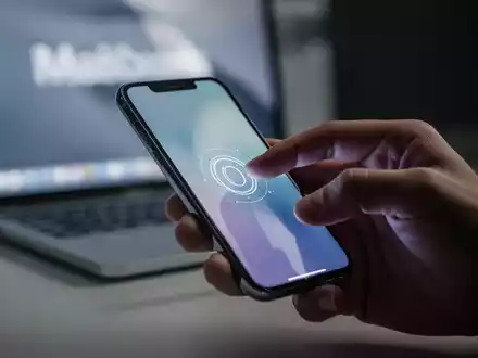 A hand tapping an iPhone screen, with a MacBook in the blurred background, illustrating wireless unlocking technology. - Foto: über boerse-global.de