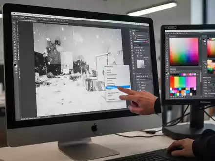 iMac Pro screen showing a washed-out image, an Eizo monitor next to it with correct colors, and a hand about to click a 'Preferences' menu item in Photoshop. - Foto: über boerse-global.de
