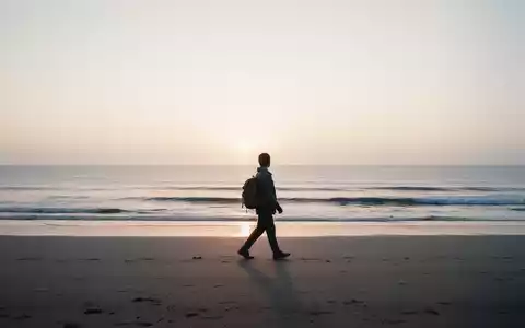 Eine einzelne Person mit kleinem Rucksack geht bei Sonnenaufgang an einem einsamen Strand entlang, mit weitem Meer im Hintergrund. - Foto: über boerse-global.de