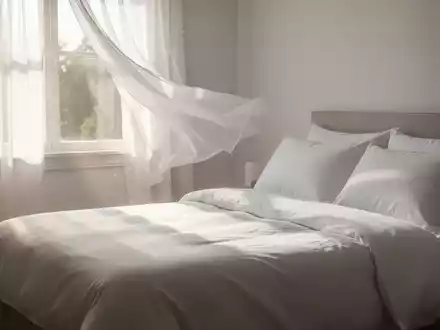 Ein von Sonnenlicht durchflutetes Schlafzimmer mit einem frisch aussehenden Bett, das mit weißen Bettdecken und Kissen bezogen ist. - Foto: über boerse-global.de