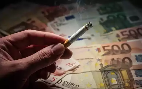 Nahaufnahme einer Hand, die eine Zigarette hält, mit unscharfen Euro-Banknoten im Hintergrund, die steigende Kosten symbolisieren. - Foto: über boerse-global.de