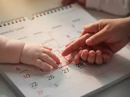 Ein Kalenderblatt mit hervorgehobenem Datum, die Hand eines Babys berührt sanft die Hand eines Erwachsenen, symbolisiert Elternzeit und Schutz. - Foto: über boerse-global.de