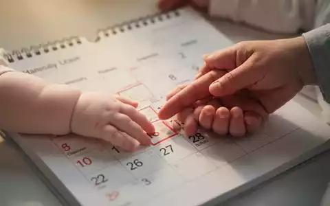 Ein Kalenderblatt mit hervorgehobenem Datum, die Hand eines Babys berührt sanft die Hand eines Erwachsenen, symbolisiert Elternzeit und Schutz. - Foto: über boerse-global.de