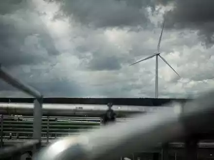 Eine einzelne Windkraftanlage vor einem turbulenten Himmel, ihre Rotorblätter stehen still. Im Vordergrund verschwommene Industrieelemente. - Foto: über boerse-global.de