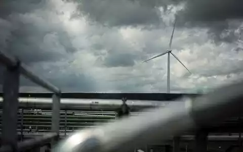 Eine einzelne Windkraftanlage vor einem turbulenten Himmel, ihre Rotorblätter stehen still. Im Vordergrund verschwommene Industrieelemente. - Foto: über boerse-global.de