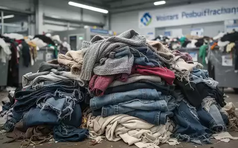 Ein Stapel bunter Altkleider und Textilien, unscharf im Hintergrund eine moderne Textilrecyclinganlage oder Sammelstelle. - Foto: über boerse-global.de