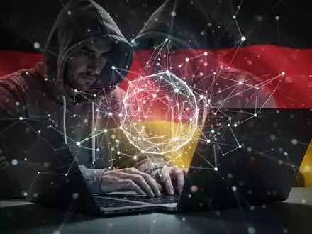 Eine schattenhafte Person mit Kapuze tippt auf einem Laptop, umgeben von einem leuchtenden, abstrakten digitalen Netzwerk, das KI-Cyberbedrohungen darstellt. - Foto: über boerse-global.de