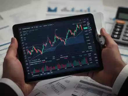 Nahaufnahme einer Hand, die ein Tablet mit Börsencharts hält, im Hintergrund unscharfe Finanzdokumente und ein Taschenrechner. - Foto: über boerse-global.de