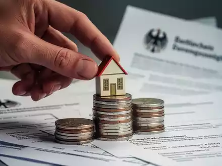 Eine Hand legt ein kleines Haus-Symbol auf einen Stapel Münzen, im Hintergrund verschwommene Regierungsdokumente. - Foto: über boerse-global.de