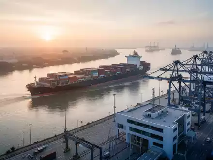 Ein großes Containerschiff auf einem Fluss bei Sonnenaufgang, mit einem modernen Hafen im Vordergrund, der effiziente Logistik symbolisiert. - Foto: über boerse-global.de