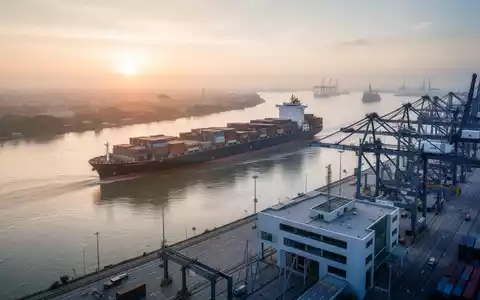 Ein großes Containerschiff auf einem Fluss bei Sonnenaufgang, mit einem modernen Hafen im Vordergrund, der effiziente Logistik symbolisiert. - Foto: über boerse-global.de