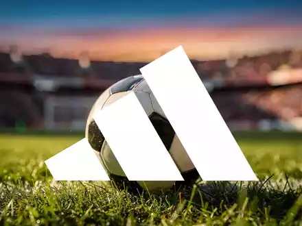 Ein Fußball mit Adidas-Logo auf einem grünen Feld, im Hintergrund ein verschwommenes Stadion und eine jubelnde Menschenmenge. - Foto: über boerse-global.de