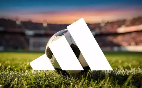 Ein Fußball mit Adidas-Logo auf einem grünen Feld, im Hintergrund ein verschwommenes Stadion und eine jubelnde Menschenmenge. - Foto: über boerse-global.de