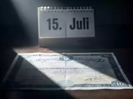 Ein Börsenzertifikat im Schatten, dahinter ein Kalenderblatt mit dem Datum '15. Juli', symbolisiert finanzielle Unsicherheit. - Foto: über boerse-global.de