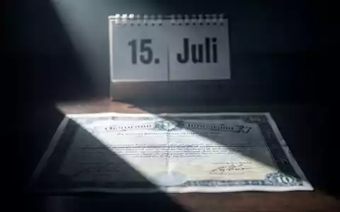 Ein Börsenzertifikat im Schatten, dahinter ein Kalenderblatt mit dem Datum '15. Juli', symbolisiert finanzielle Unsicherheit. - Foto: über boerse-global.de