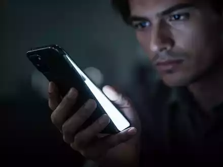 Eine Hand hält ein leuchtendes Smartphone in einer verschwommenen, dunklen Umgebung, die digitale Ablenkung symbolisiert. - Foto: über boerse-global.de
