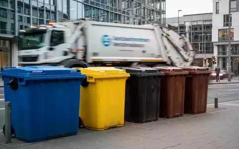 Eine Reihe von Mülltonnen in verschiedenen Farben steht auf einem Bürgersteig in einer deutschen Stadt, mit einem Müllwagen im Hintergrund. - Foto: über boerse-global.de