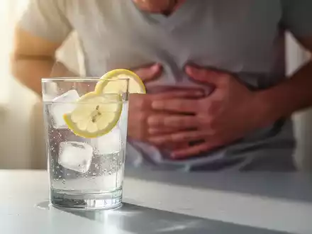 Ein Glas Wasser mit Zitrone auf einem Tisch; im verschwommenen Hintergrund hält sich eine Person leicht den Bauch. Symbolisiert Hydrierung und Magenprobleme. - Foto: über boerse-global.de