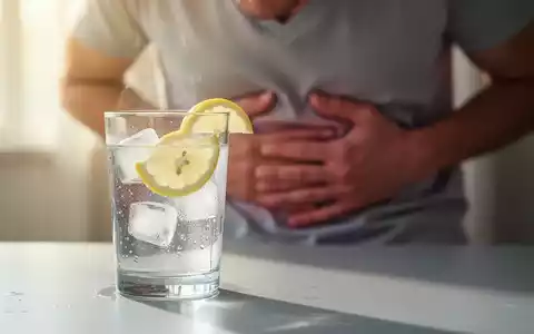 Ein Glas Wasser mit Zitrone auf einem Tisch; im verschwommenen Hintergrund hält sich eine Person leicht den Bauch. Symbolisiert Hydrierung und Magenprobleme. - Foto: über boerse-global.de