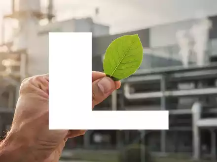 Eine Hand hält ein grünes Blatt vor einem unscharfen Fabrikhintergrund, symbolisiert Nachhaltigkeit und Wachstum für Lenzing. - Foto: über boerse-global.de