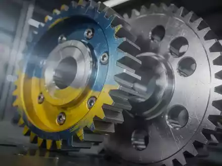 Zwei ineinandergreifende Zahnräder, eines in ukrainischen Farben, vor einem verschwommenen industriellen Hintergrund, symbolisieren Technologie-Kooperation. - Foto: über boerse-global.de