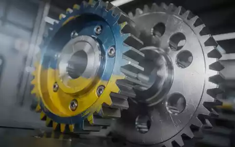 Zwei ineinandergreifende Zahnräder, eines in ukrainischen Farben, vor einem verschwommenen industriellen Hintergrund, symbolisieren Technologie-Kooperation. - Foto: über boerse-global.de