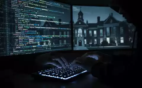 A hacker's hands typing on a keyboard in a dark room, with code and university buildings on a screen, symbolizing a cyber-attack. - Foto: über boerse-global.de
