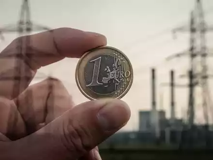 Eine Hand hält eine einzelne Euro-Münze mit verschwommenen Stromleitungen und einem Kraftwerk im Hintergrund. - Foto: über boerse-global.de