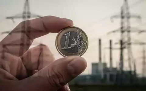 Eine Hand hält eine einzelne Euro-Münze mit verschwommenen Stromleitungen und einem Kraftwerk im Hintergrund. - Foto: über boerse-global.de