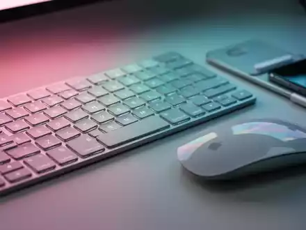 Apple Magic Keyboard and Mouse with a thin MagSafe grip, bathed in soft pink, teal, and blue light. - Foto: über boerse-global.de