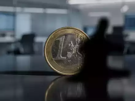 Eine einzelne stilisierte Euromünze, teilweise von einem Schatten verdeckt, auf einer reflektierenden Oberfläche in einem Finanzbüro. - Foto: über boerse-global.de