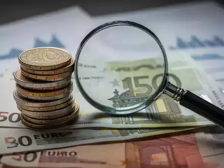 Ein Stapel deutscher Euro-Münzen und -Banknoten mit einer Lupe, die auf Finanzdokumenten oder Diagrammen liegt. - Foto: über boerse-global.de