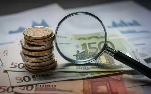Ein Stapel deutscher Euro-Münzen und -Banknoten mit einer Lupe, die auf Finanzdokumenten oder Diagrammen liegt. - Foto: über boerse-global.de