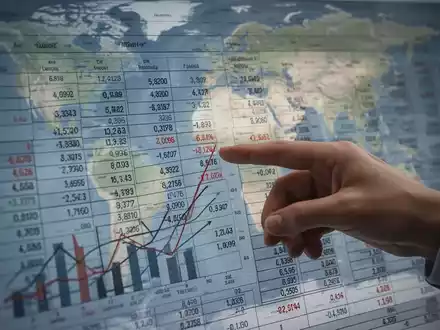 Eine Hand zeigt auf einen Finanzbericht mit Zahlen und Diagrammen, im Hintergrund eine angedeutete Weltkarte. - Foto: über boerse-global.de