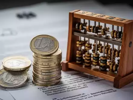 Ein Stapel deutscher Euro-Münzen neben einem Abakus, mit verschwommenen Dokumenten im Hintergrund, symbolisiert finanzielle Berechnungen und Beiträge. - Foto: über boerse-global.de