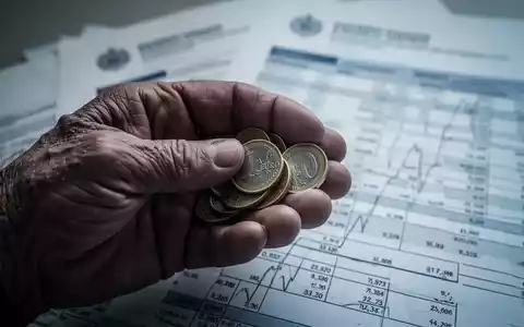 Eine Nahaufnahme einer älteren Hand, die ein paar Euro-Münzen zählt, mit verschwommenen offiziellen Dokumenten im Hintergrund. - Foto: über boerse-global.de