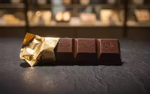 Eine teilweise ausgepackte Lindt-Schokoladentafel liegt auf einer dunklen Oberfläche, die ihre luxuriöse Textur hervorhebt. - Foto: über boerse-global.de