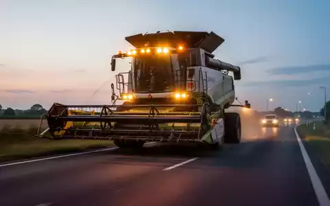 Ein überbreiter Mähdrescher mit Warnleuchten fährt bei Dämmerung auf einer Landstraße. - Foto: über boerse-global.de