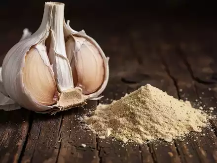 Nahaufnahme einer halbierten frischen Knoblauchknolle neben einem kleinen Haufen Knoblauchpulver auf dunklem Holz. - Foto: über boerse-global.de