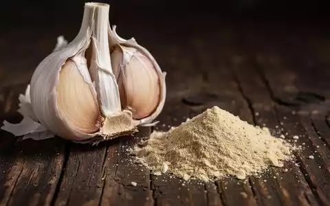 Nahaufnahme einer halbierten frischen Knoblauchknolle neben einem kleinen Haufen Knoblauchpulver auf dunklem Holz. - Foto: über boerse-global.de