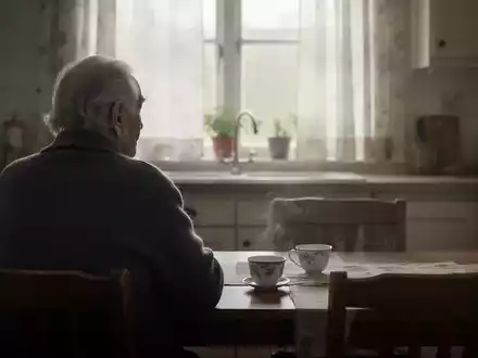 Eine ältere Person sitzt allein am Küchentisch und blickt nachdenklich aus dem Fenster. Eine Tasse steht auf dem Tisch. - Foto: über boerse-global.de