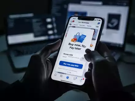 A hand holding a smartphone showing a shopping app with a 'buy now, pay later' option, against a blurred, dimly lit background with a laptop. - Foto: über boerse-global.de