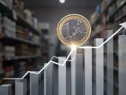 Eine einzelne Euro-Münze balanciert auf einem stilisierten, stabilen Balkendiagramm, mit verschwommenen Konsumgütern im Hintergrund. - Foto: über boerse-global.de