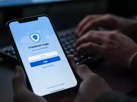 A close-up of a smartphone screen showing a fake O2 login page, with blurred hands typing on a keyboard in the background. - Foto: über boerse-global.de